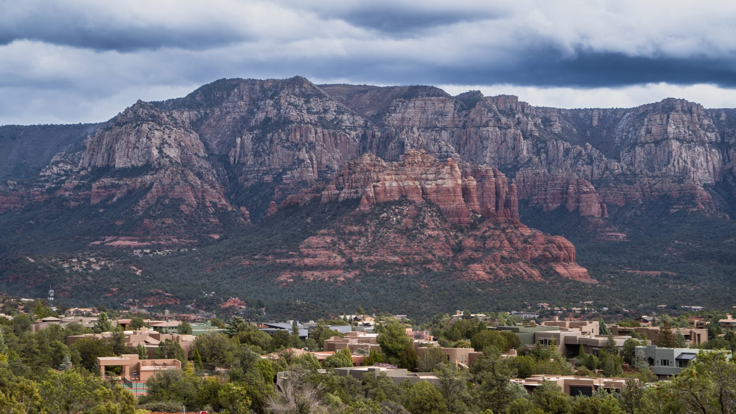View of Sedona's red rock formations with a residential area at its base, under a cloudy sky.
