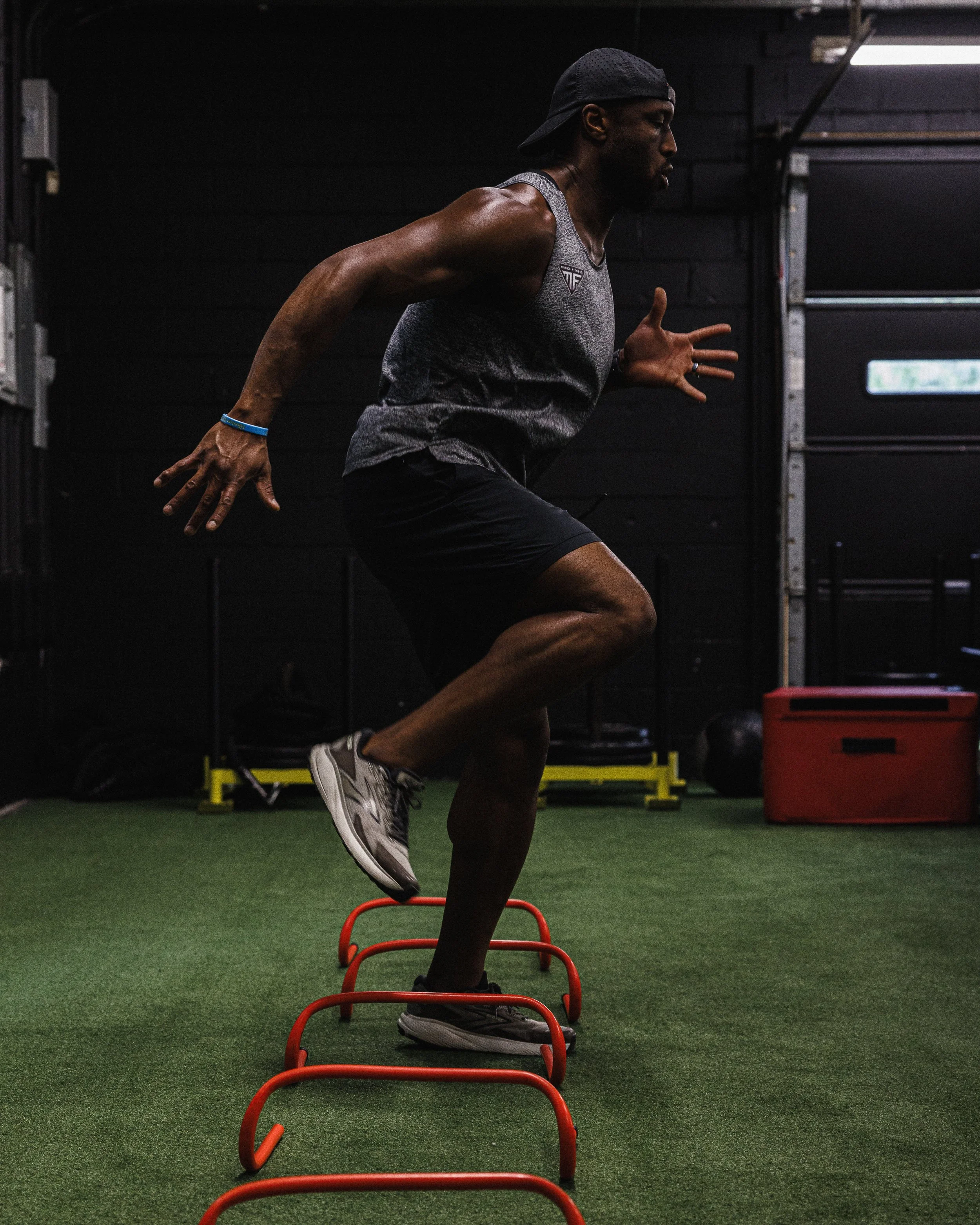Man in a gray sleeveless shirt, black shorts, and gray sneakers doing agility drills with red hurdles in a gym.