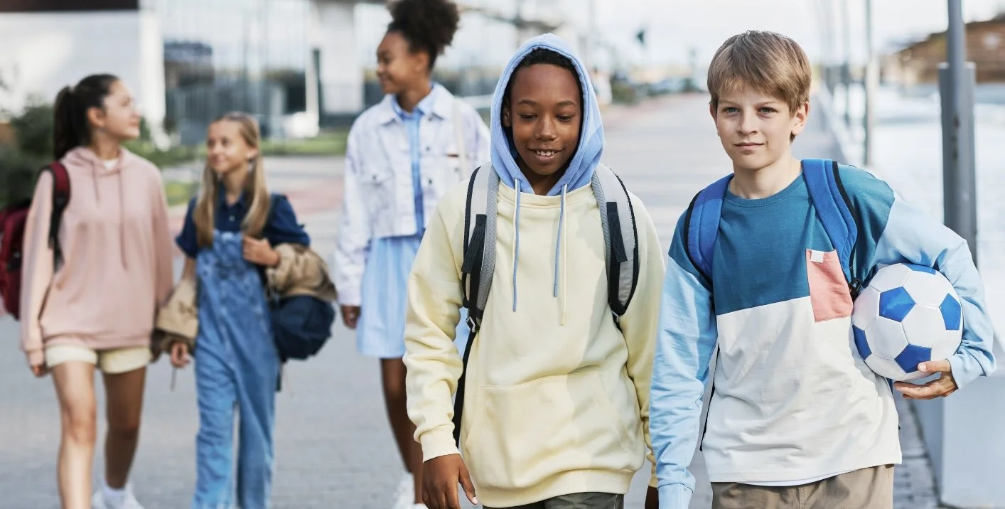 Group of five children walking outdoors, carrying backpacks and a soccer ball, with a modern building in the background.