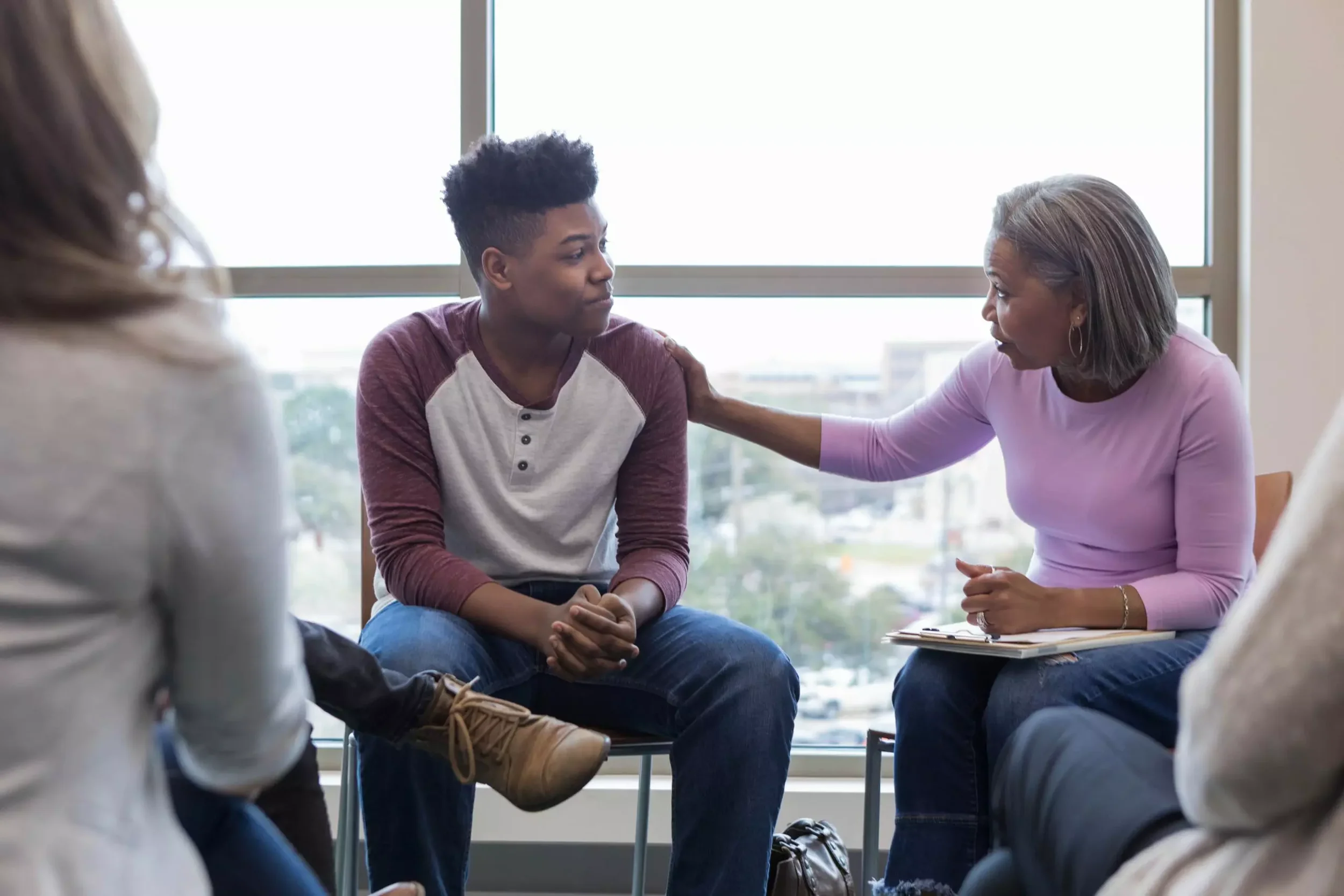 A young man sits in an office or counseling room, receiving supportive contact from an older woman, possibly a counselor, who is gently holding his arm, in a comforting gesture. Other people are paying attention in the background.