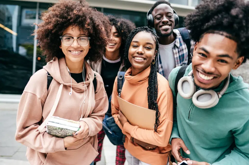 Group of five diverse smiling young adults standing outdoors in front of a building, wearing casual clothing and backpacks.