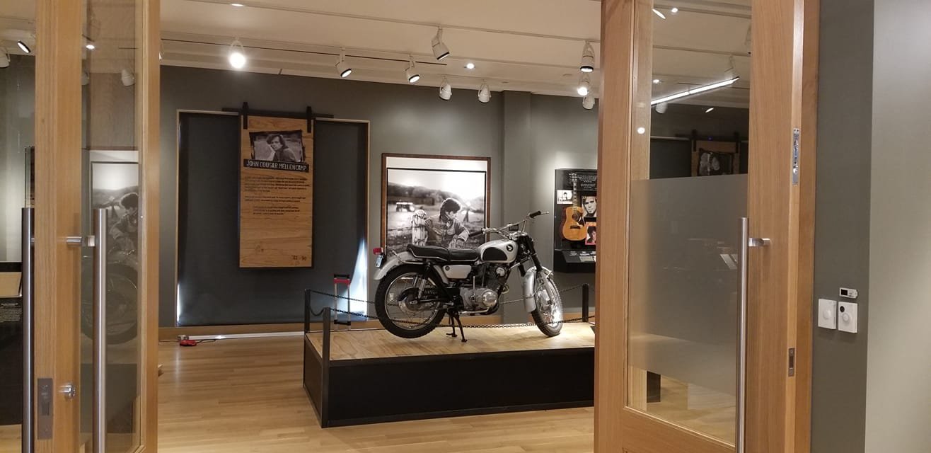 Vintage motorcycle displayed on a raised wooden platform in a museum exhibit, with informational panels and black-and-white photographs in the background.