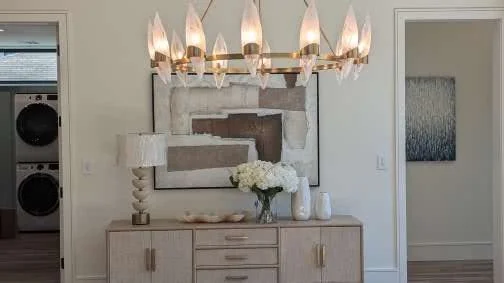A modern sideboard with a vase of white flowers, a table lamp, and decorative objects, in a living room with abstract artwork and a chandelier.