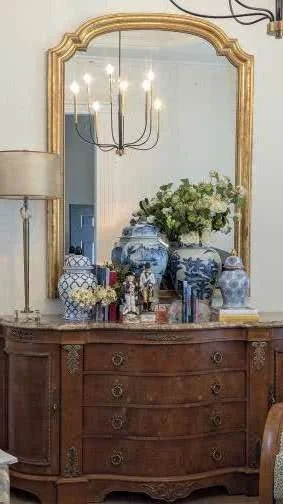 A wooden dresser with a large mirror and decorative items including blue and white vases and a bouquet of flowers, with a chandelier hanging above.