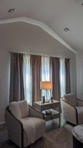 Living room with two beige armchairs, a side table with a lamp, and curtains covering large windows.