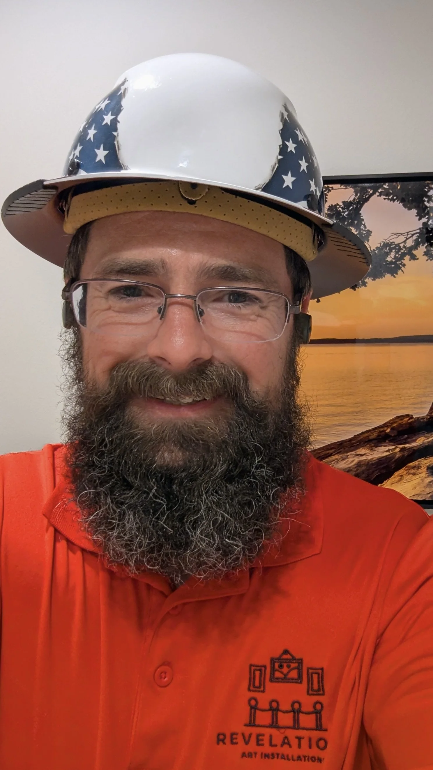 A man with a beard and glasses wearing an orange shirt and a star-spangled helmet, smiling in front of a scenic background of a sunset over a lake and trees.