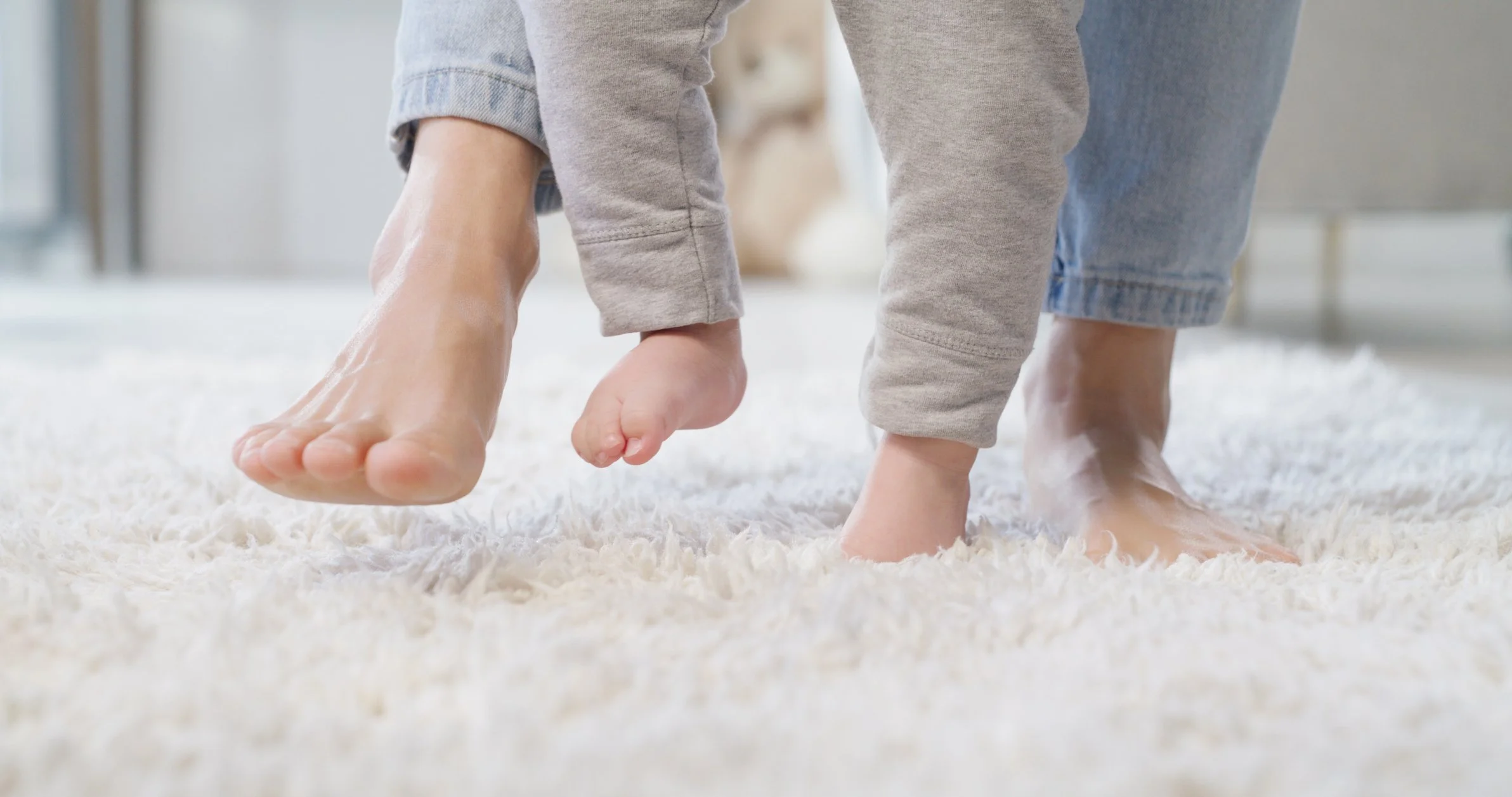 A person helping a small child walk on a white rug indoors.