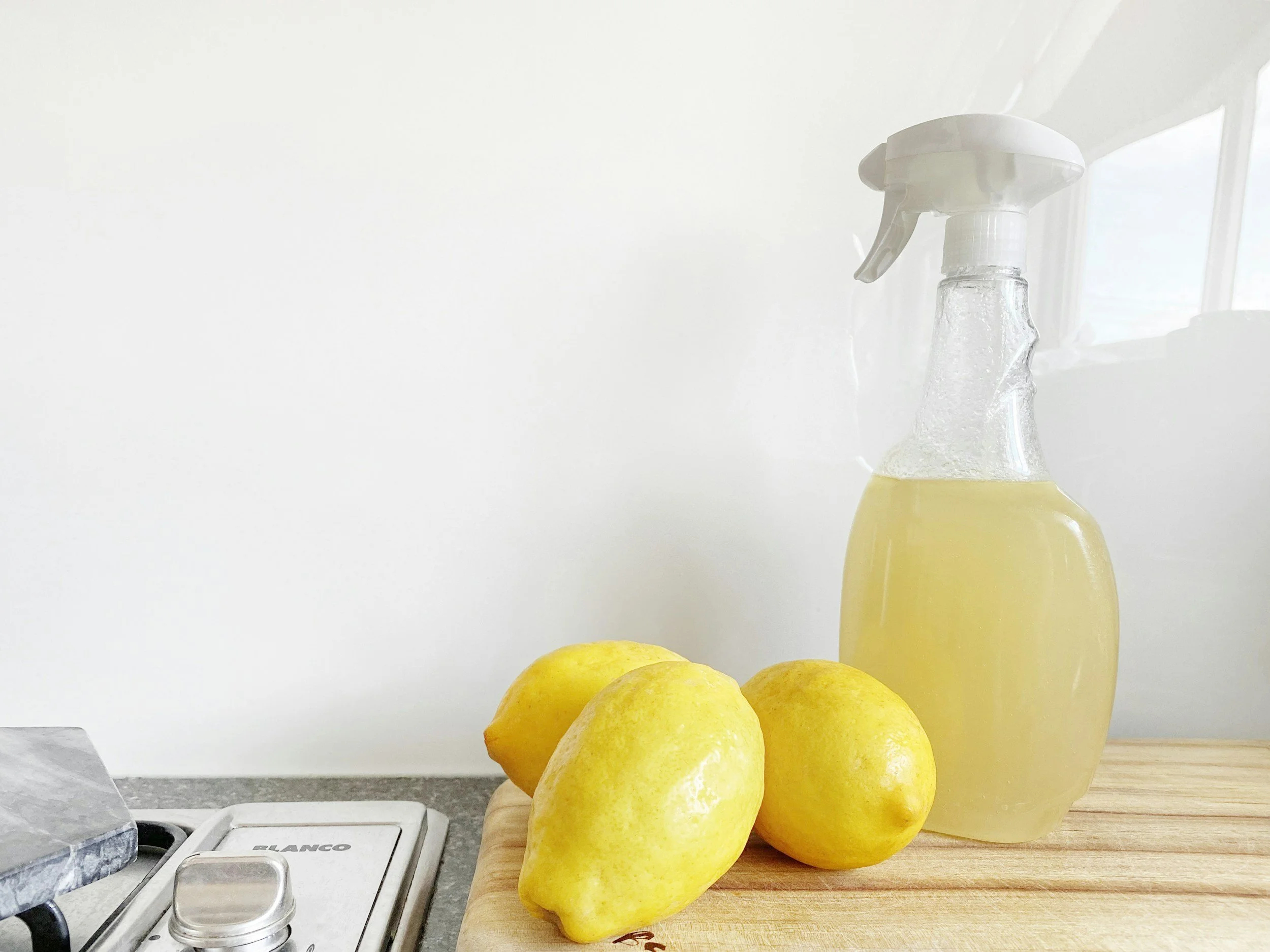 Three yellow lemons and a spray bottle filled with lemon cleaner on a wooden cutting board against a white background.