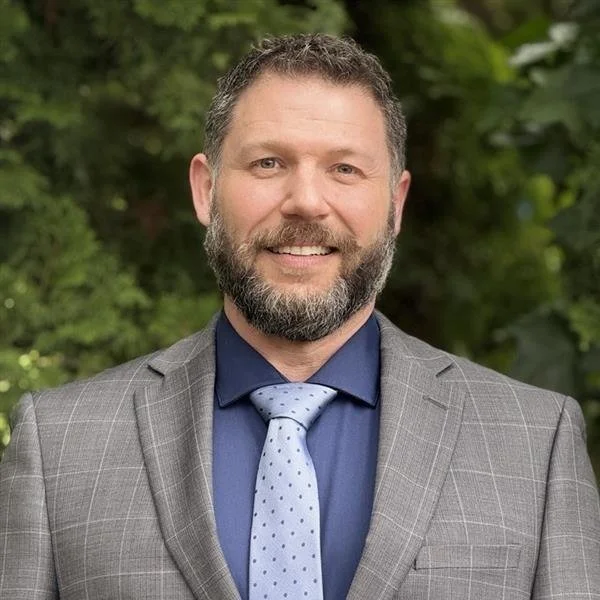 Portrait of a smiling man with a beard, wearing a red collared shirt against a gray background.