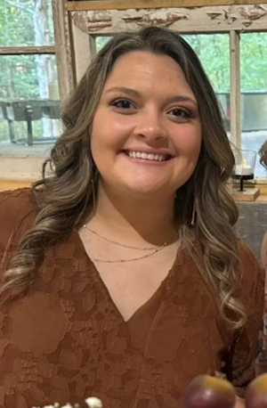 A woman smiling indoors with wooden walls and large windows in the background.