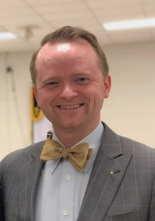 A smiling man in a gray suit with a gold bow tie, indoors with a plain ceiling and blurred background.
