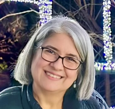 A woman with gray hair and glasses smiling outdoors at night, with illuminated trees in the background.