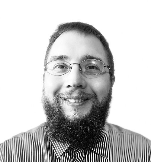 A smiling man with glasses and a beard, wearing a striped collared shirt, against a plain light background.