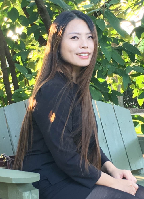 Smiling woman with long brown hair sitting on a green outdoor bench surrounded by leafy trees.