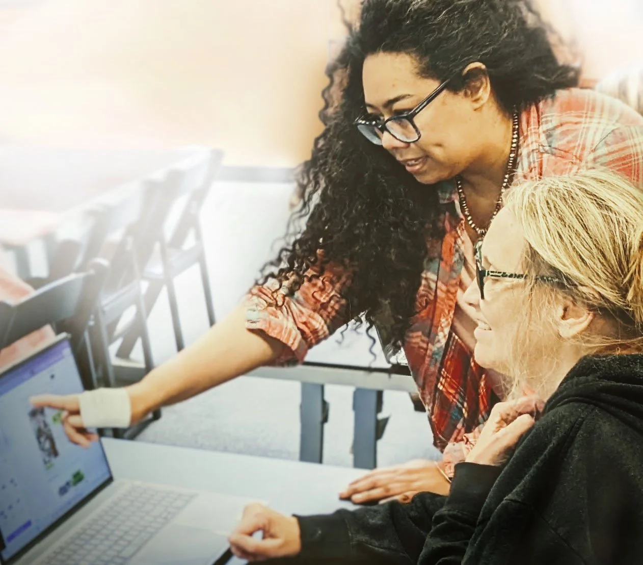 Two women, one a teacher with dark curly hair and glasses, and the other with blonde hair and glasses, look at a laptop screen together in a classroom setting.