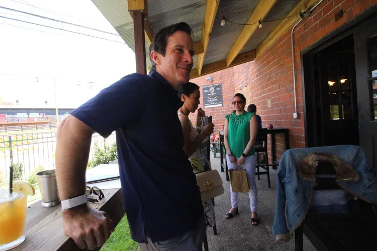 Group of people socializing outdoors on a patio at a restaurant or bar, with a man in a blue shirt smiling in the foreground, two women and a young man in the background, one woman holding a drink, and others engaged in conversation.