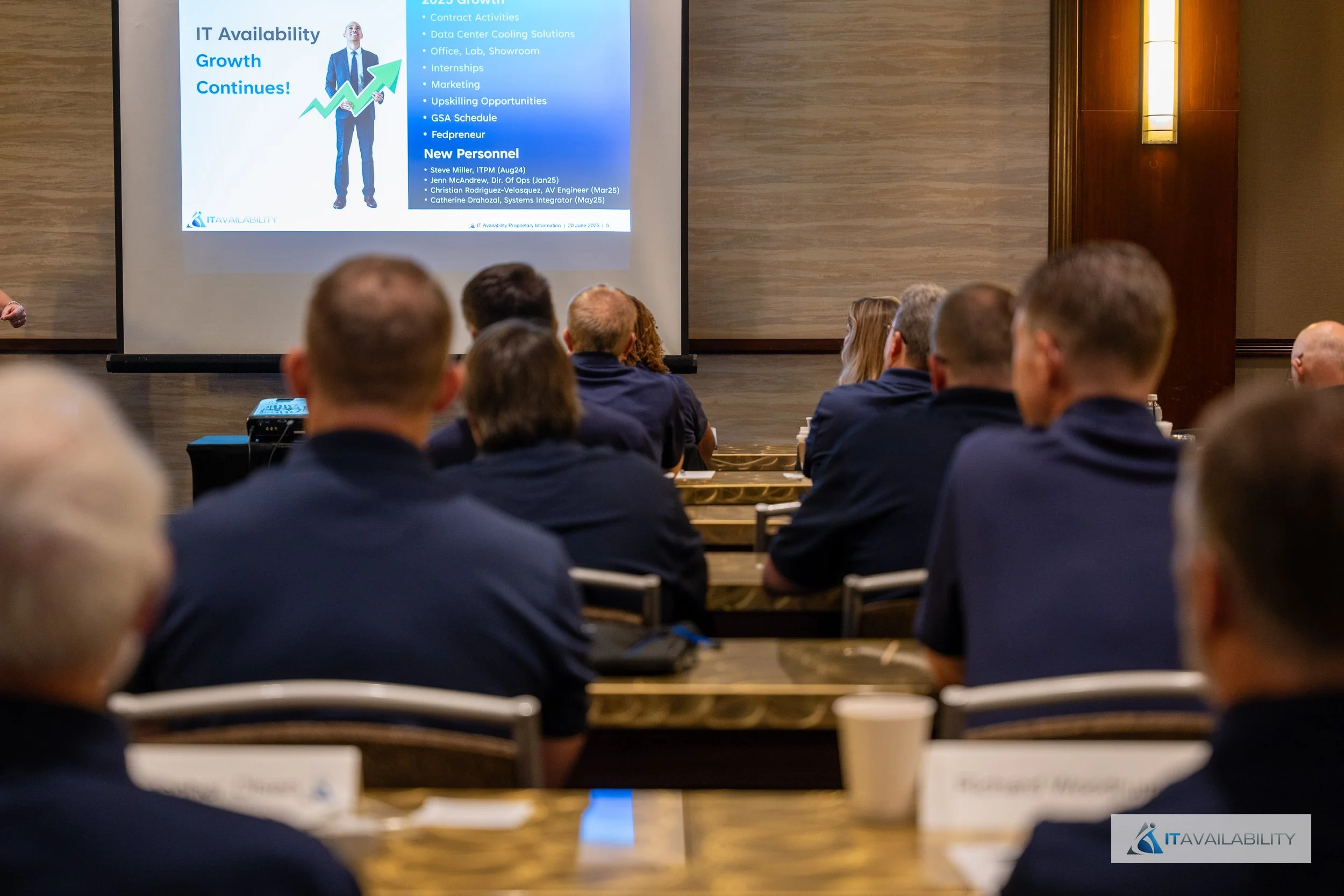 A group of people attending a presentation in a conference room with a large screen displaying a slide titled 'IT Availability Growth Continues!' and some bullet points related to IT activities and new personnel.