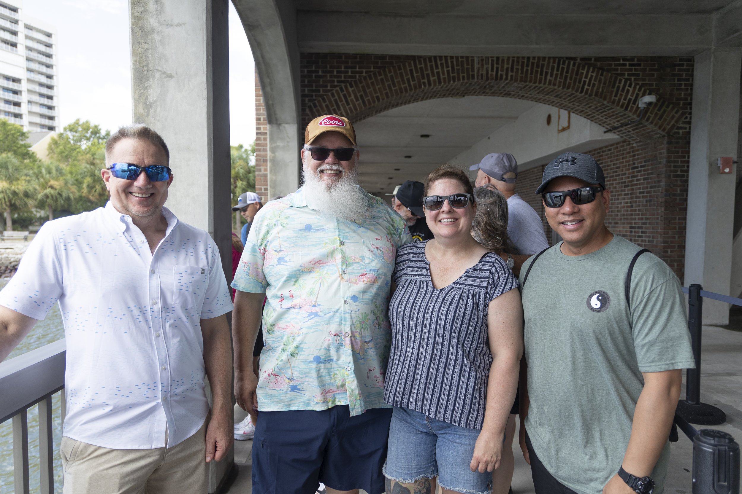 Four people smiling and posing outdoors, wearing sunglasses and casual summer clothes, with a brick building and other people in the background.