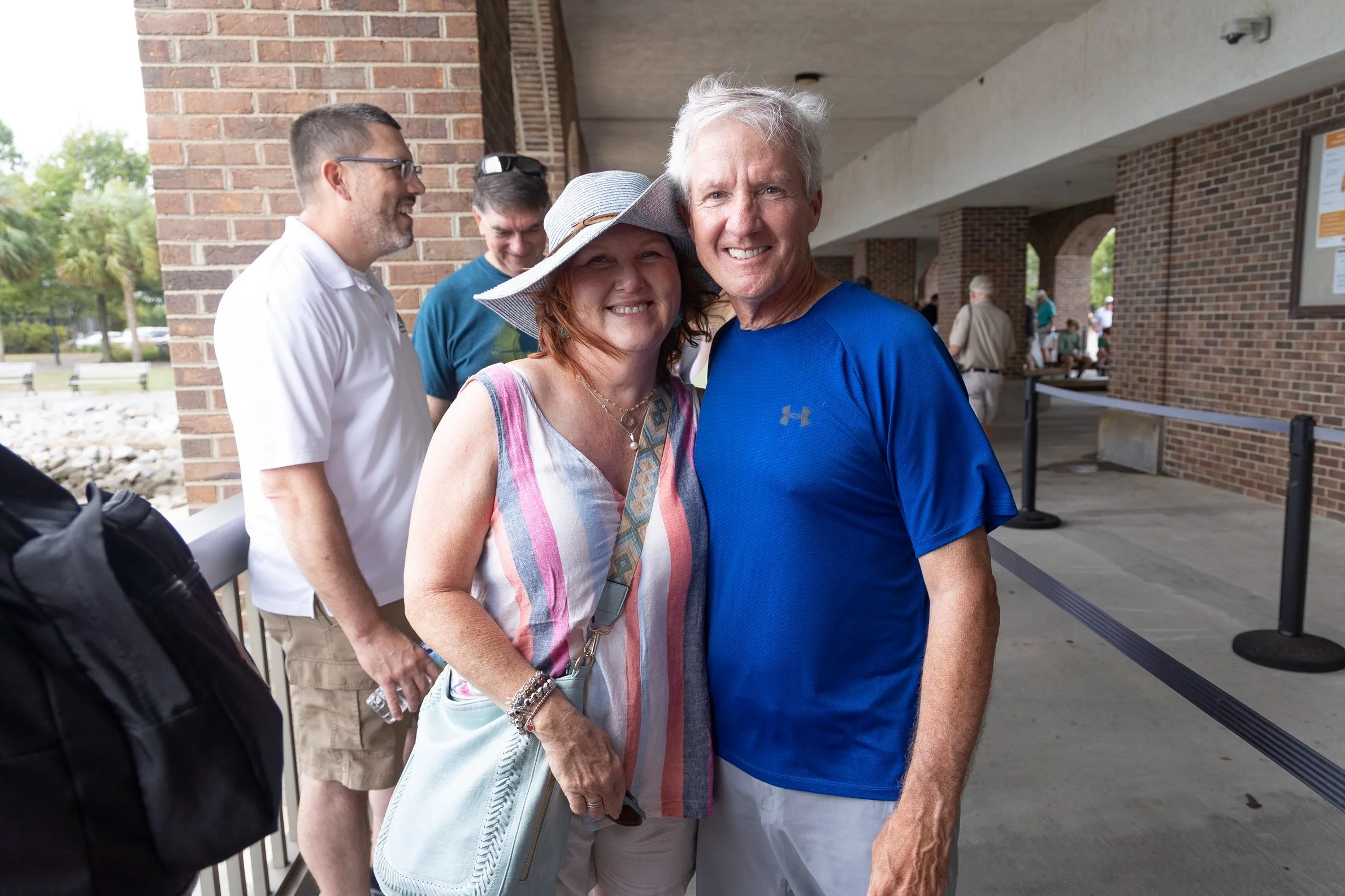 Smiling couple standing close together outdoors in front of a brick building, with other people in the background.
