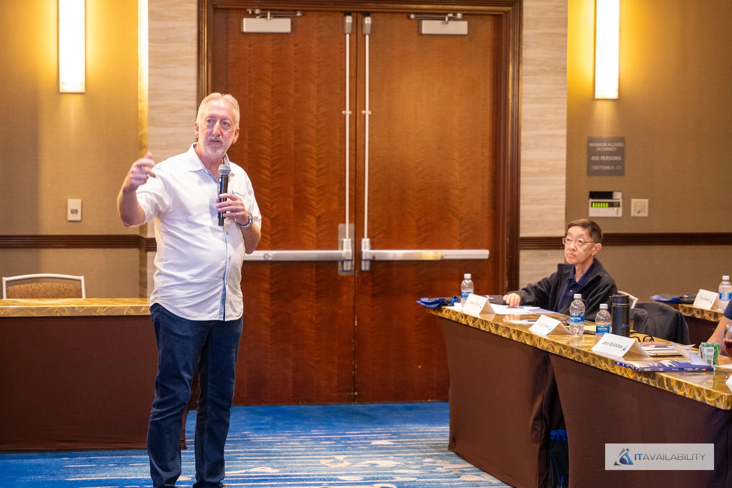 A man in a white shirt holding a microphone speaks to a conference room audience, with another man seated at a table beside him. The room has wooden doors in the background and water bottles on the tables.