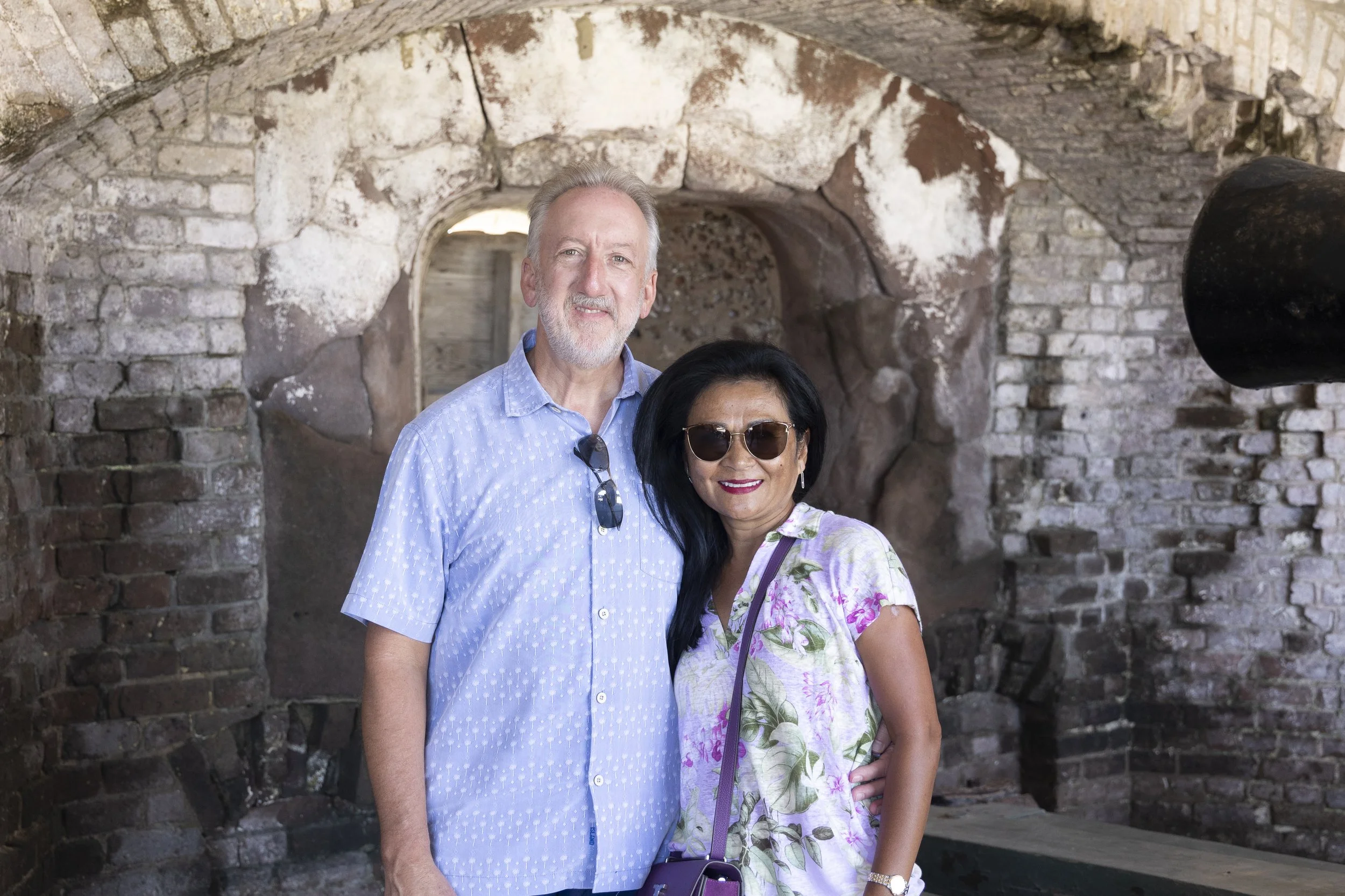 A smiling middle-aged man with a beard and a woman with dark hair, wearing sunglasses, standing close together in front of a rustic brick archway.