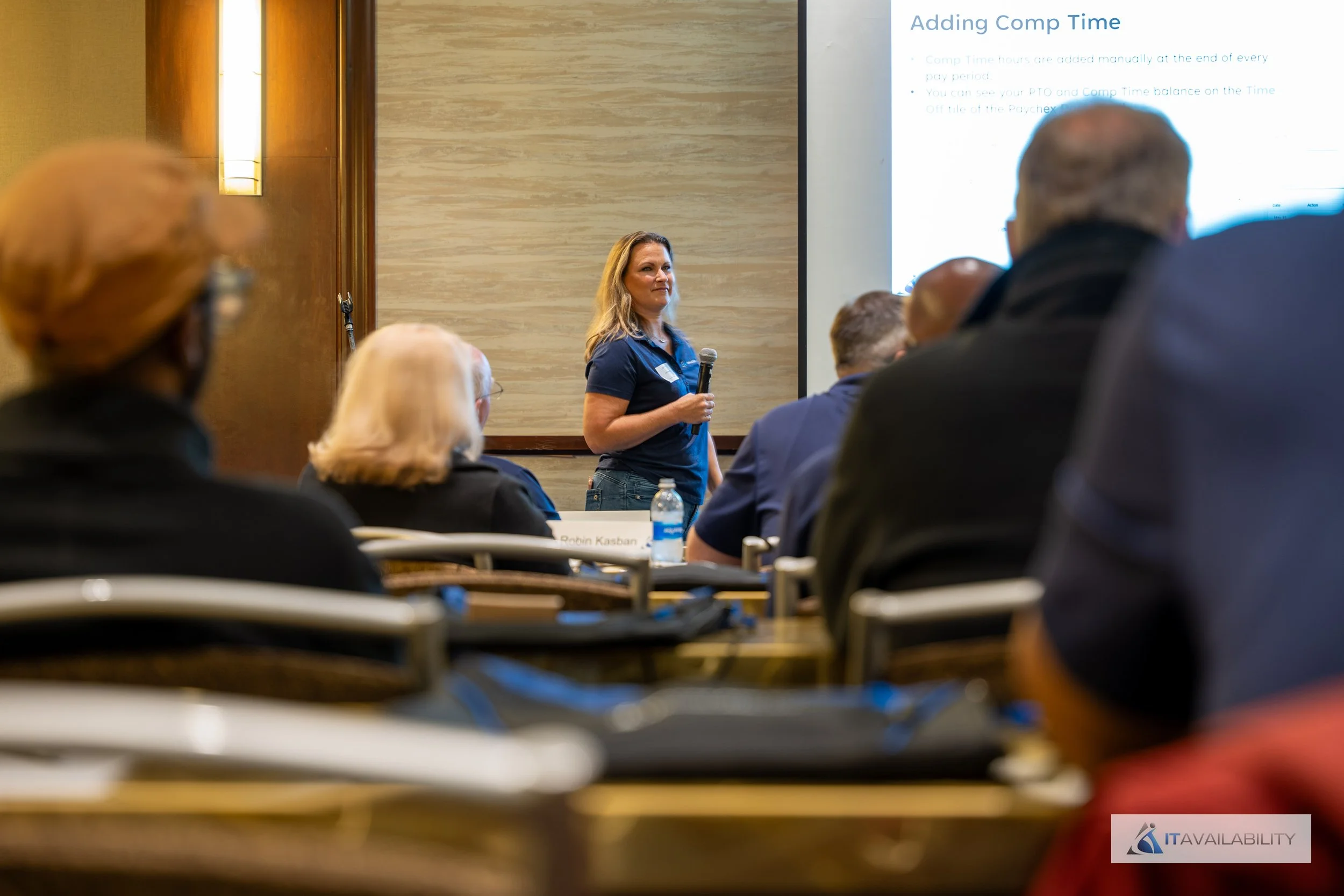 Woman giving a presentation in a conference room to an audience, with a slide titled 'Adding Comp Time' projected on the screen.