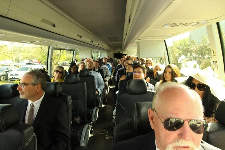 Passengers seated inside a tour bus during daytime, with some people wearing sunglasses and hats, and sunlight streaming through the windows.