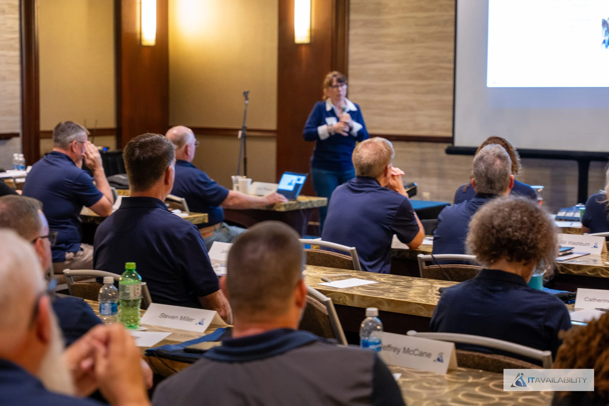 A woman presenting at a conference with attendees seated at tables, looking at her and a large screen.