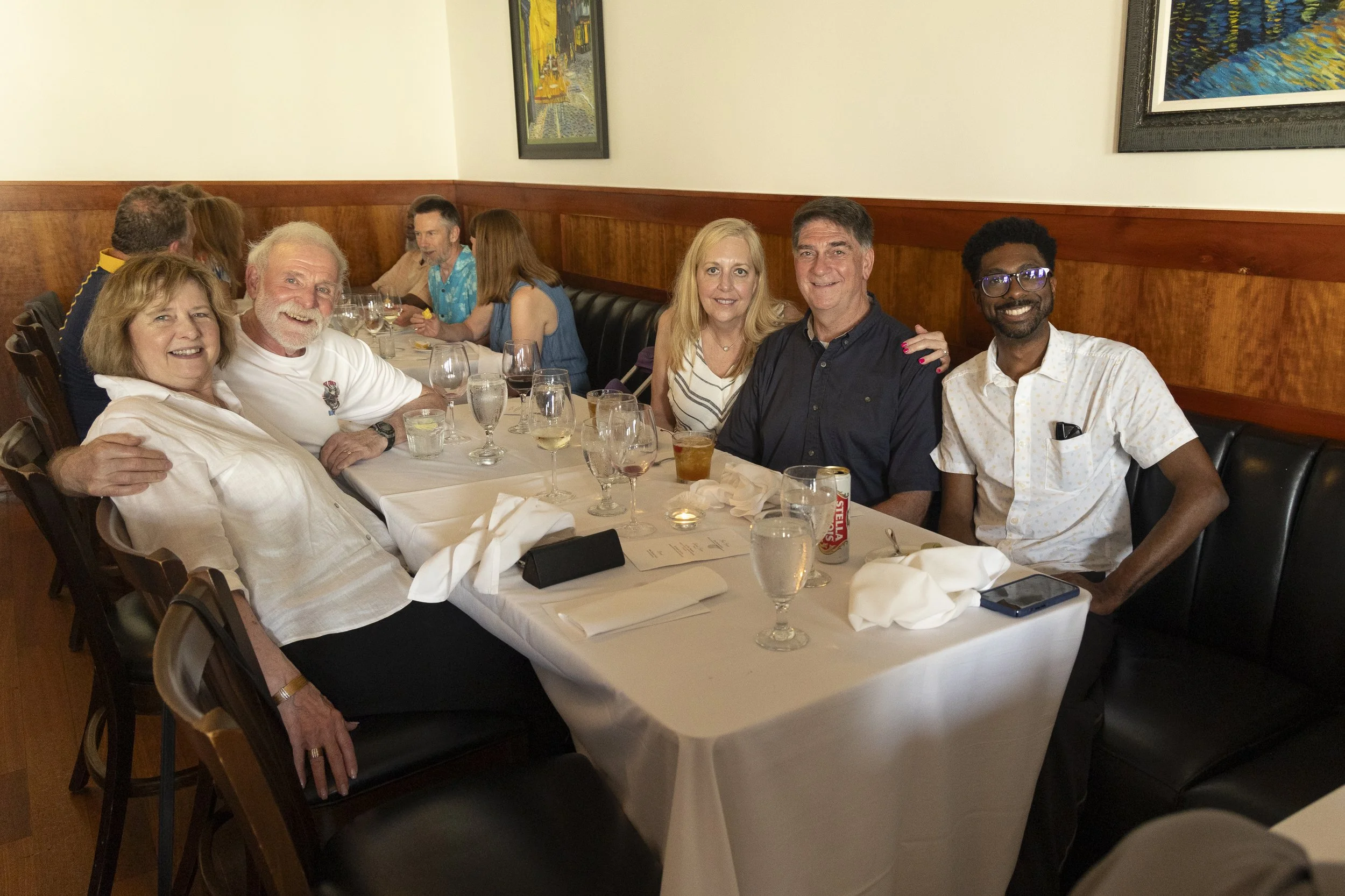A group of six people sitting at a restaurant table, smiling, with glasses of drinks and a white tablecloth, with other diners in the background.
