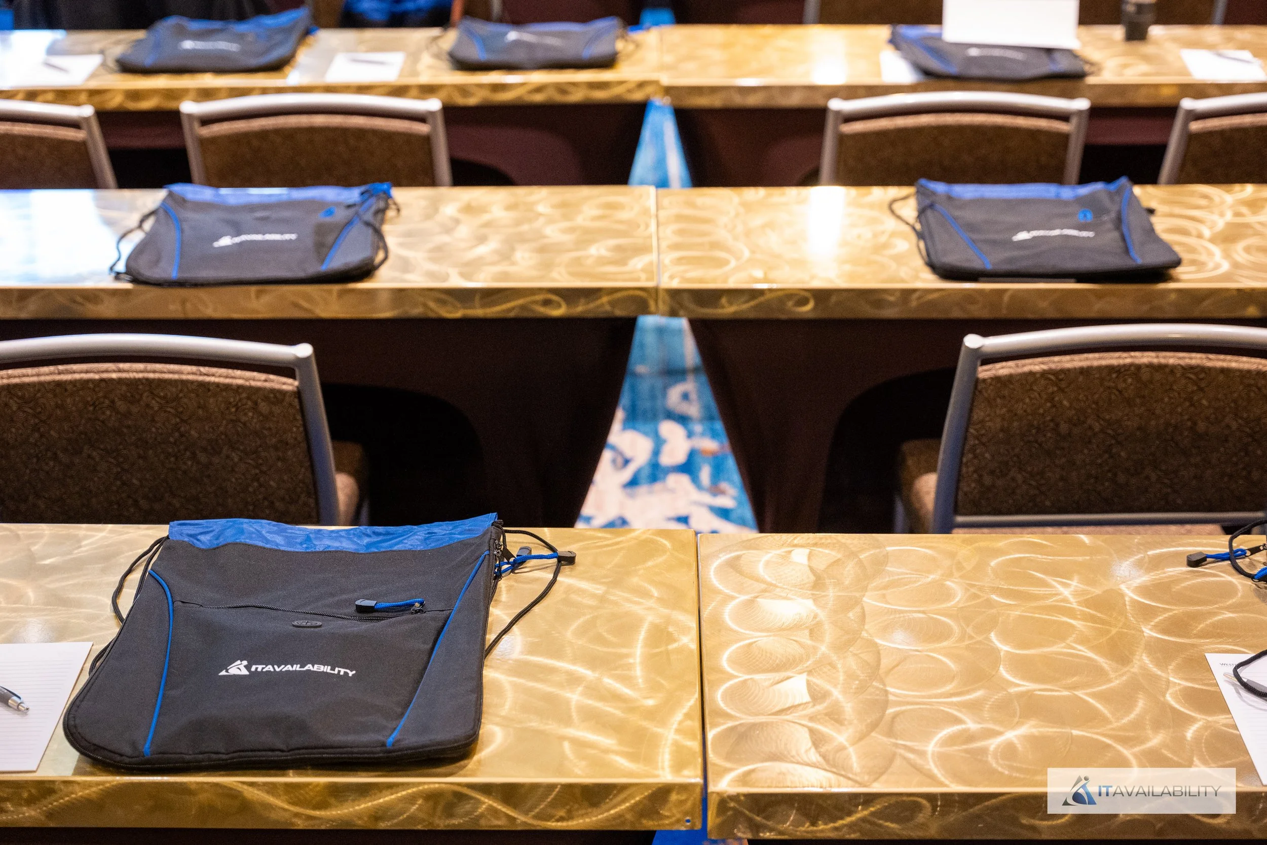 Conference room with empty tables, each with a black and blue tote bag and a piece of paper, arranged in rows.