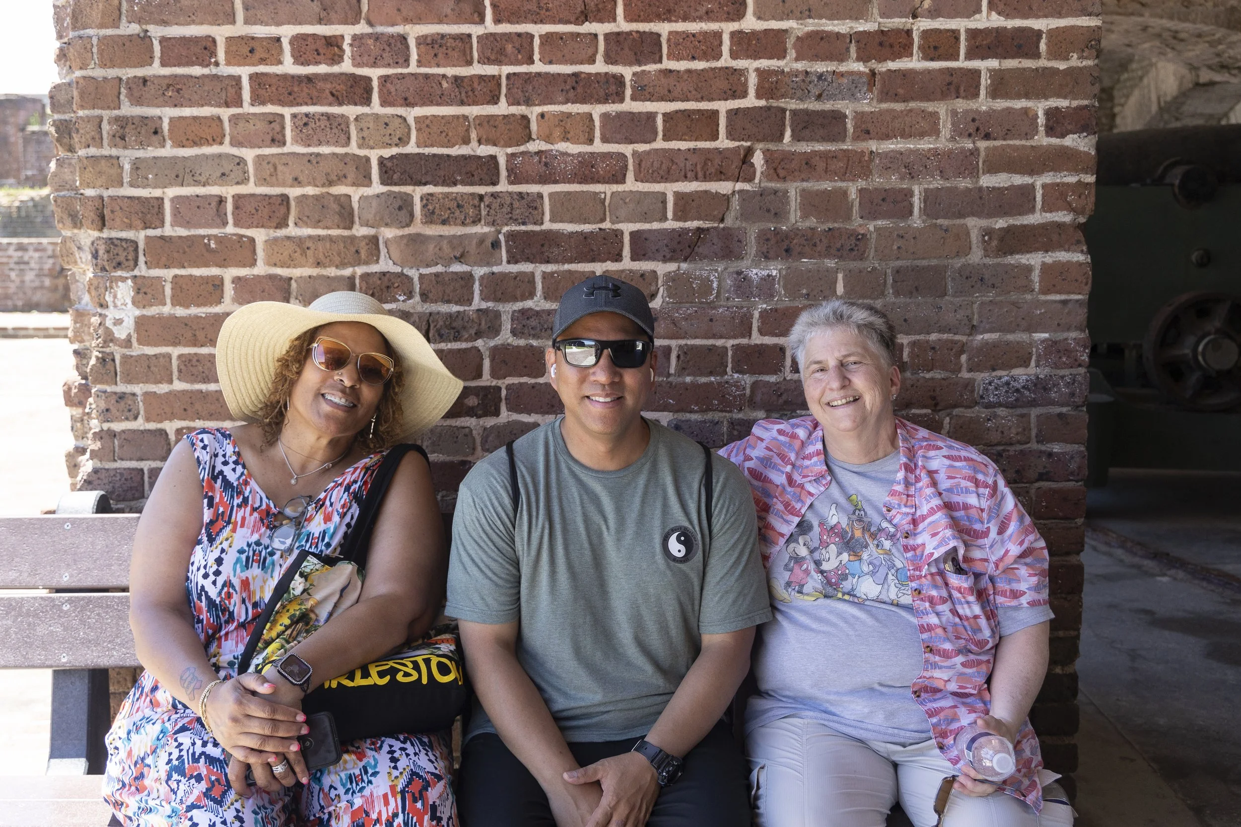 Three people sitting on a bench in front of a brick wall, smiling and relaxing. The woman on the left is wearing a wide-brimmed hat, sunglasses, and a colorful dress. The man in the middle is wearing sunglasses, a baseball cap, and a gray t-shirt. Th