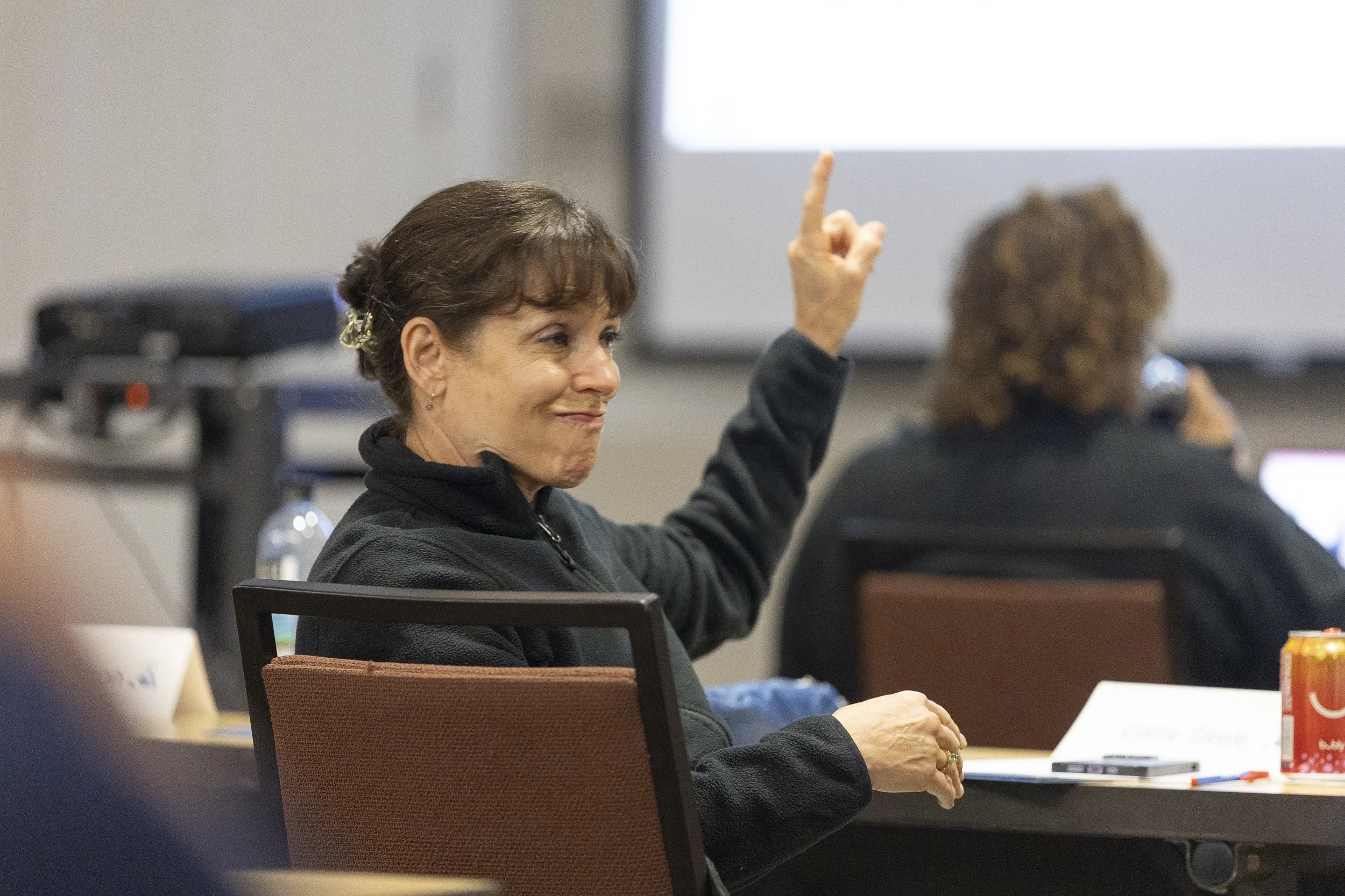 A woman with short brown hair and earrings, raising her hand in a classroom or conference setting, with a woman with curly hair and a microphone in the background.