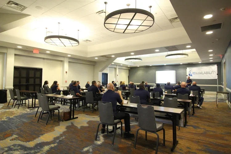 A conference room with attendees seated at tables, listening to a speaker at the front, with a presentation screen and a banner that reads "IT Availability."