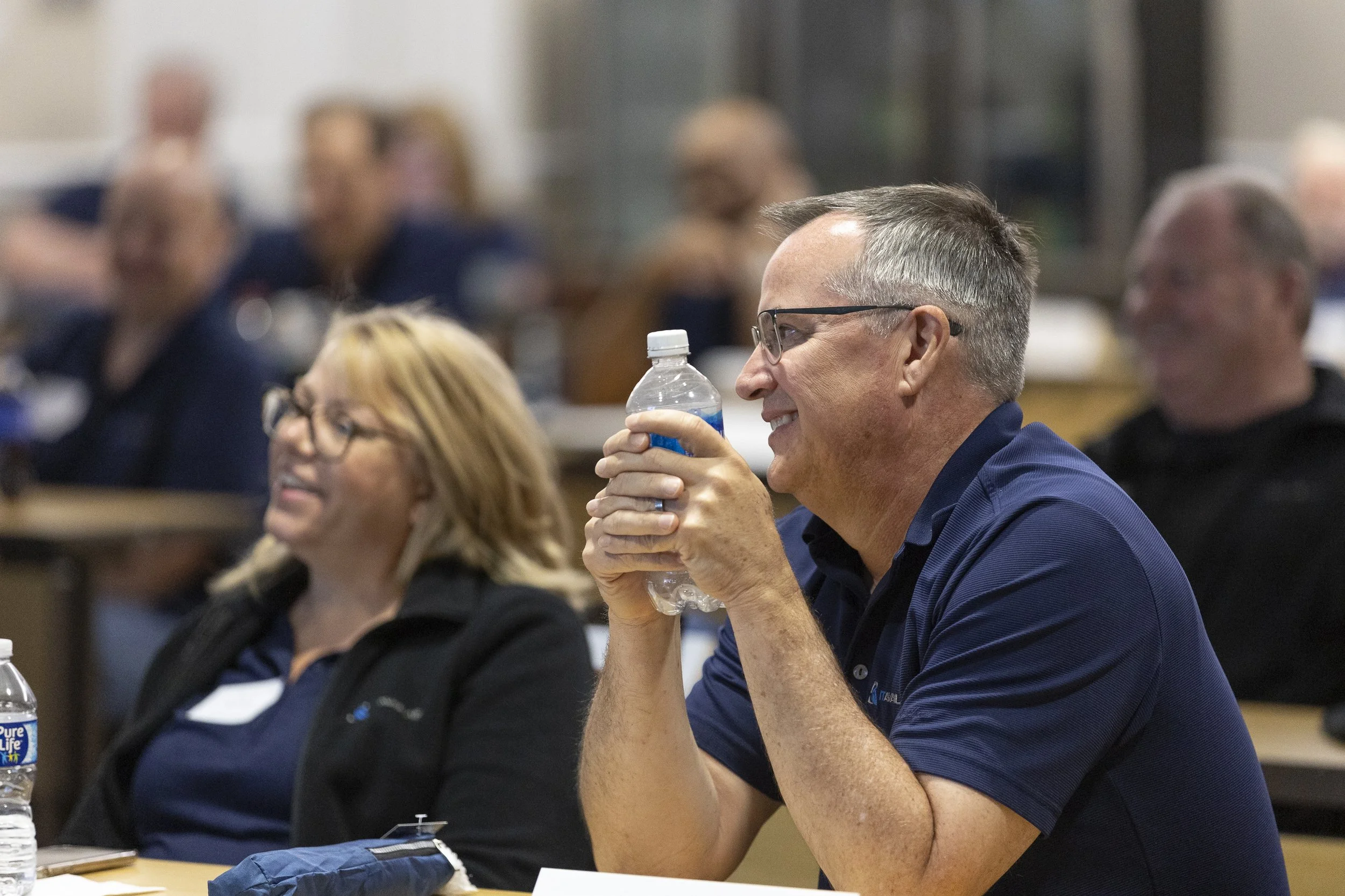 Man smiling, wearing glasses and a navy shirt, holding a water bottle during a meeting or conference, with blurred attendees in the background.