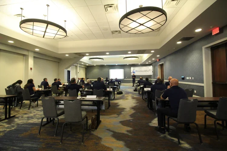 A conference room with attendees seated at tables, listening to a presenter at the front with a screen, in a professional setting.