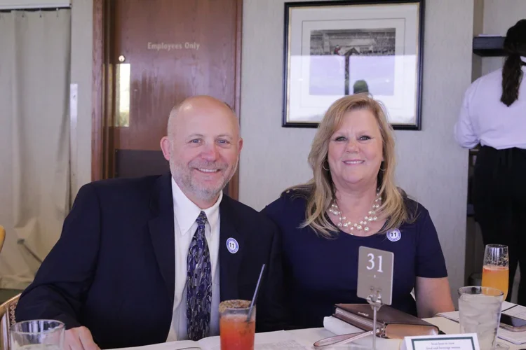 A man and woman sitting at a table during a formal event, smiling at the camera. The man wears a dark suit with a patterned tie, and the woman wears a dark dress with pearl jewelry. There are drinks and a table number on the table.