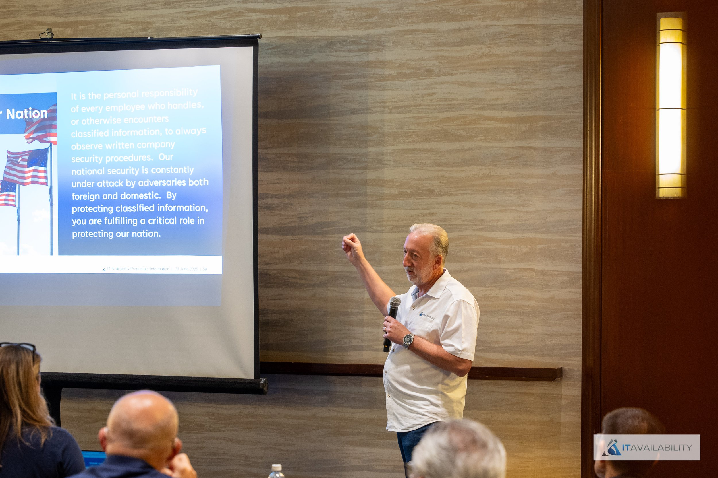 An older man with gray hair and beard giving a presentation in a conference room, holding a microphone in his right hand and gesturing with his left hand. A projector screen displays a slide about national security and employee responsibility. The au