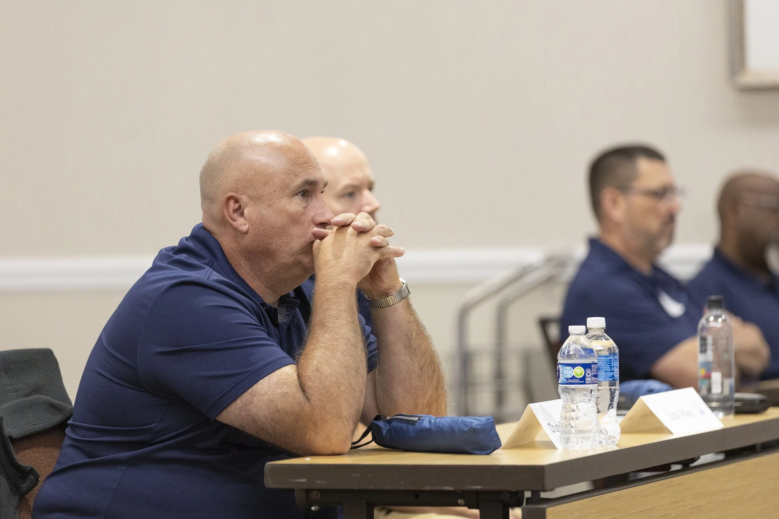 Man with a bald head sitting at a table with his hands clasped near his face, attentively listening during a meeting, with water bottles and paper name tags in front of him.