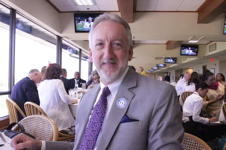 A man with gray hair and beard wearing a gray suit with a purple tie, sitting at a dining table in a busy restaurant or event space, surrounded by other people, with large windows and multiple TV screens in the background.