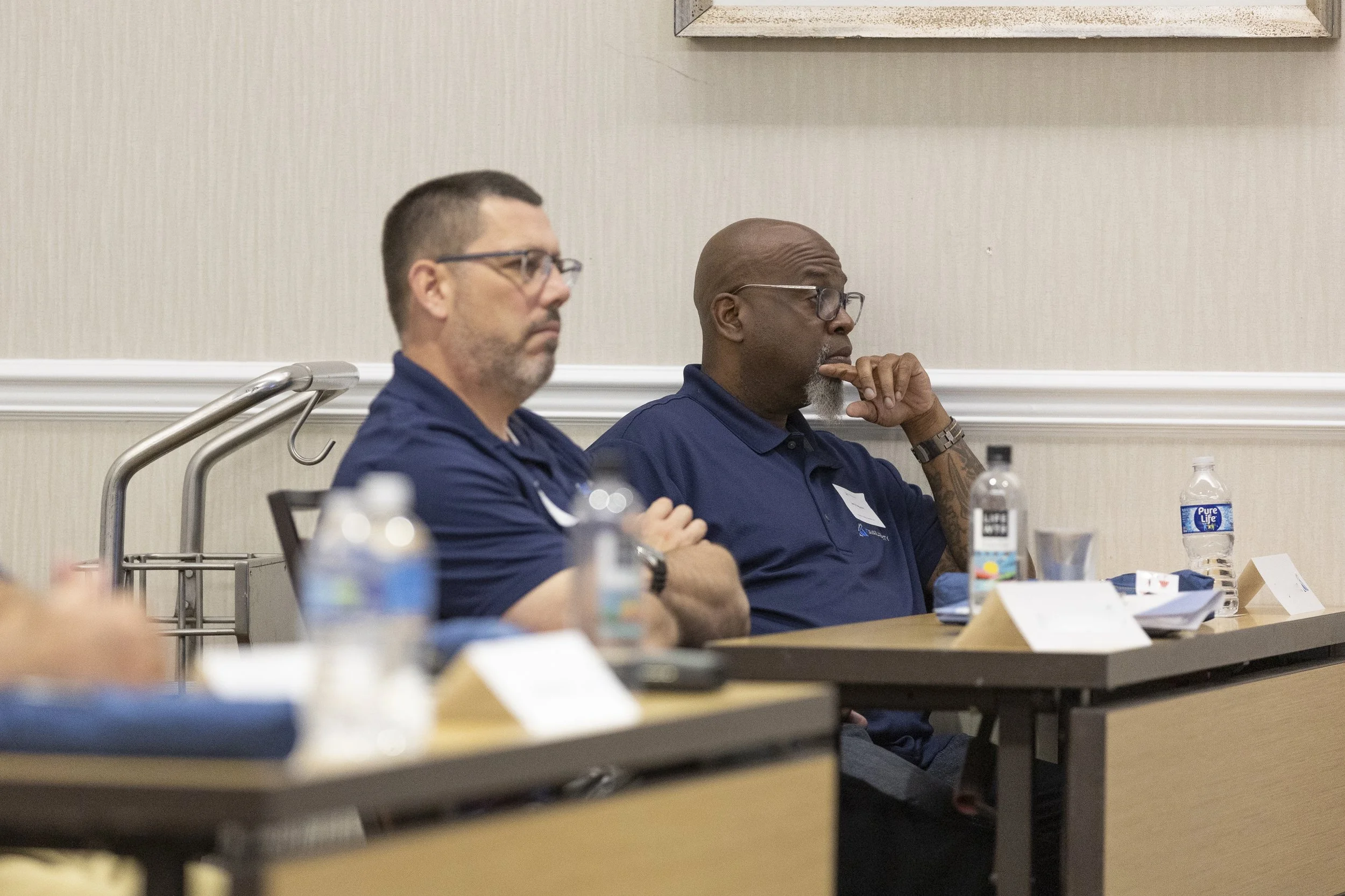 Two men sitting at a conference table, listening attentively; one is wearing glasses and a dark blue shirt, and the other is wearing glasses and a matching dark blue shirt, with a tattoo on his arm.