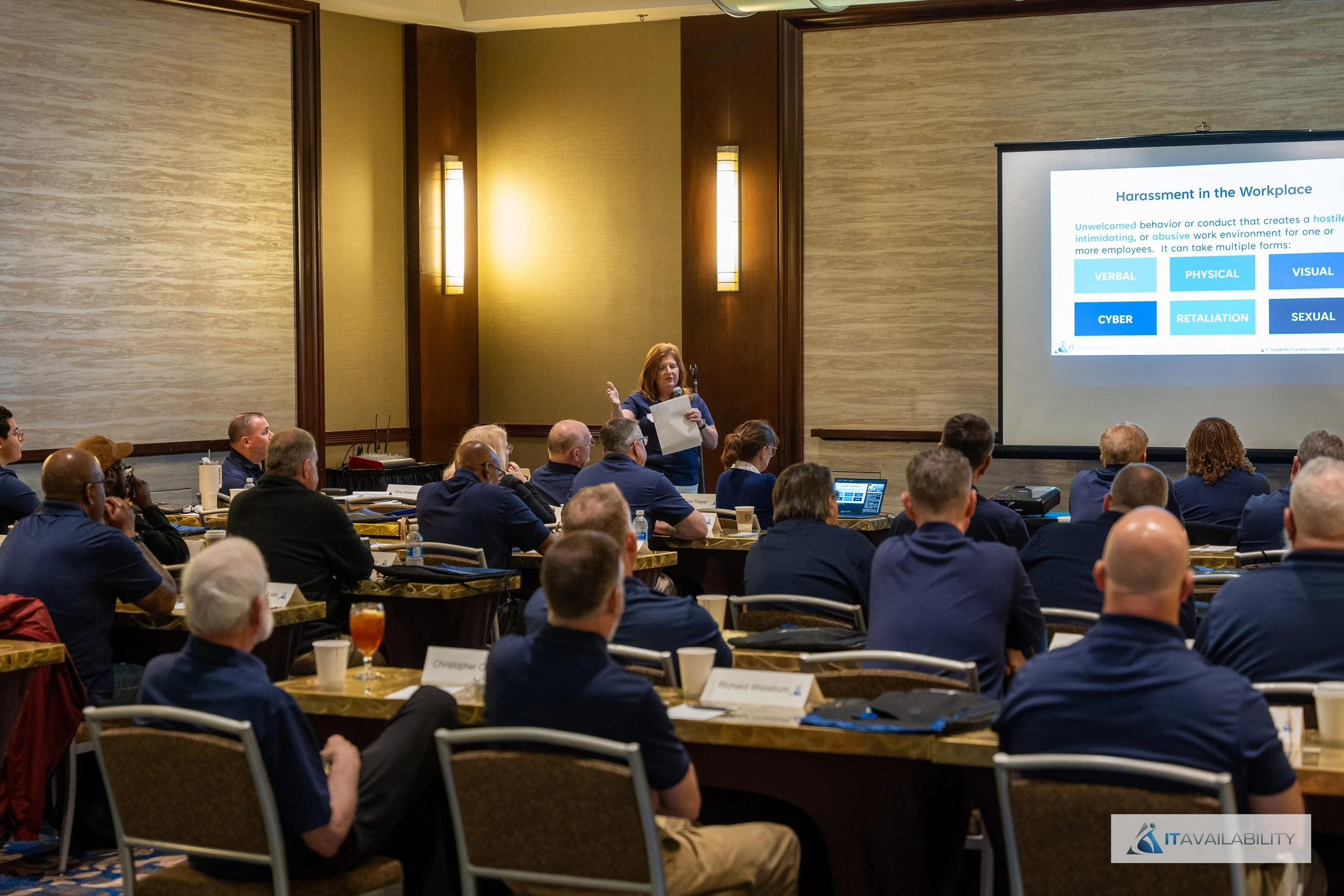 A woman giving a presentation at a conference with a slide on workplace harassment displayed on a large screen. Attendees sit at tables with notebooks, drinks, and name tags in a well-lit conference room.