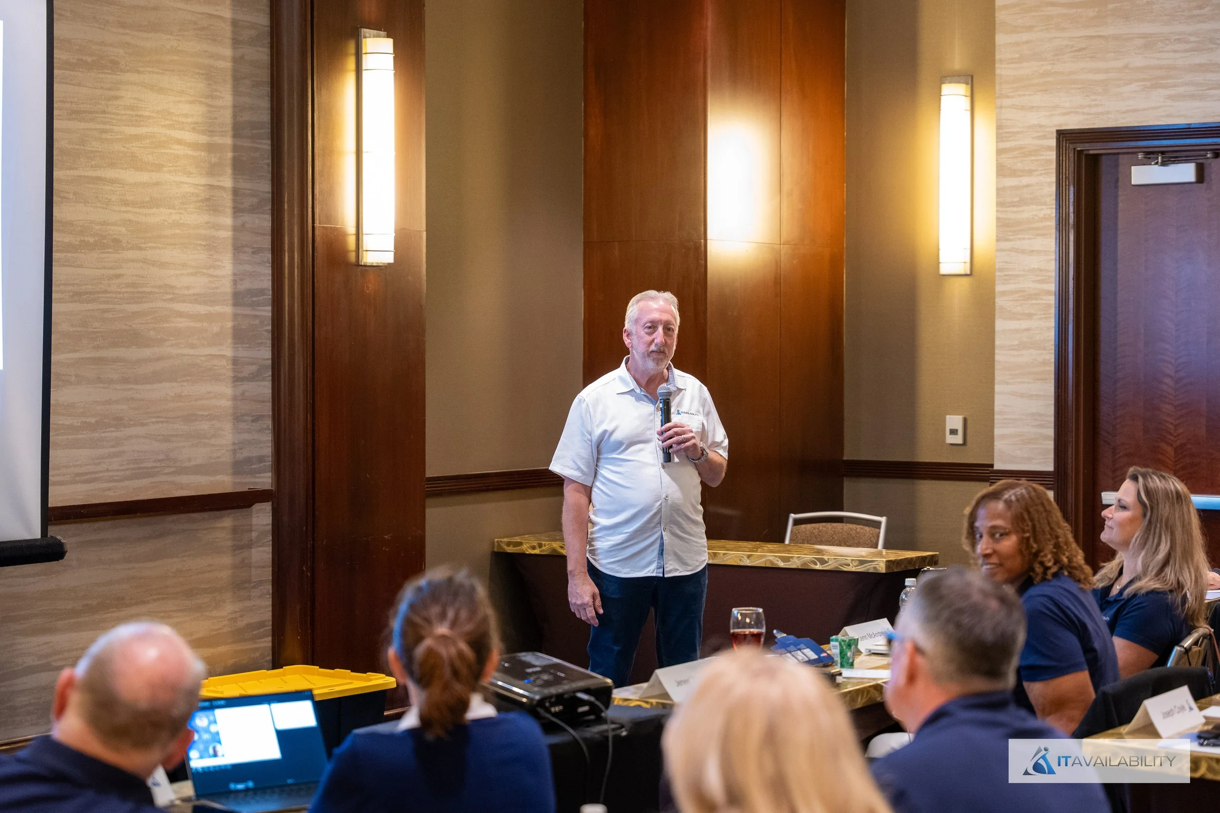 A man standing at the front of a conference room holding a microphone, giving a presentation to an audience seated at tables.
