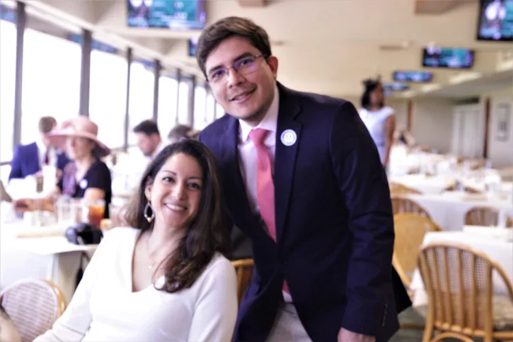 A man and woman are smiling at a restaurant or event space with multiple tables and other guests in the background.
