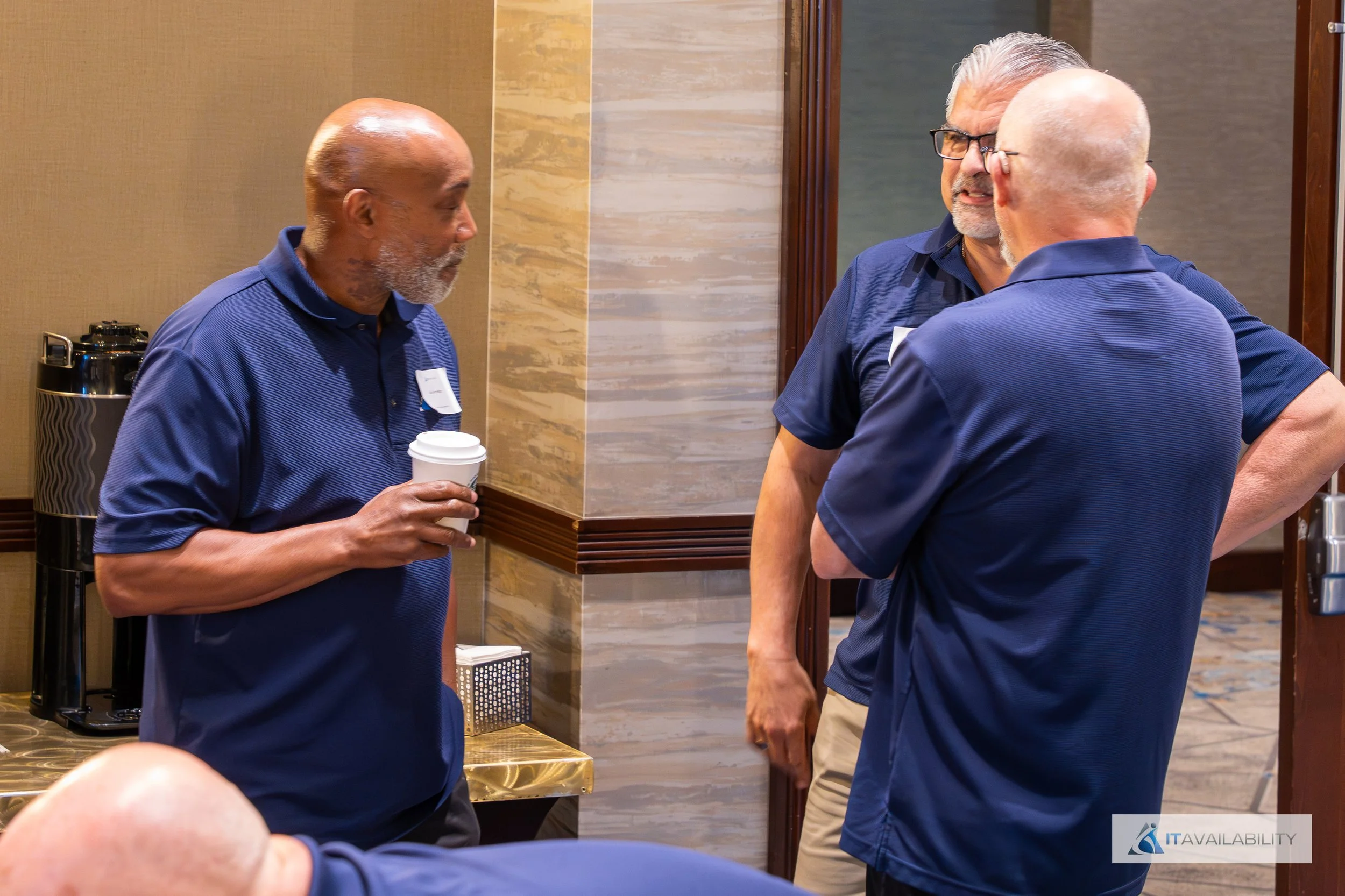 Three older men in blue shirts engaged in conversation inside a conference room, one holding a coffee cup.