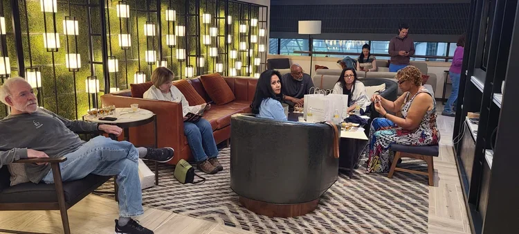 People sitting and standing in a modern lounge area with seating, tables, and decorative lighting, some using their phones and others reading or engaging with each other.