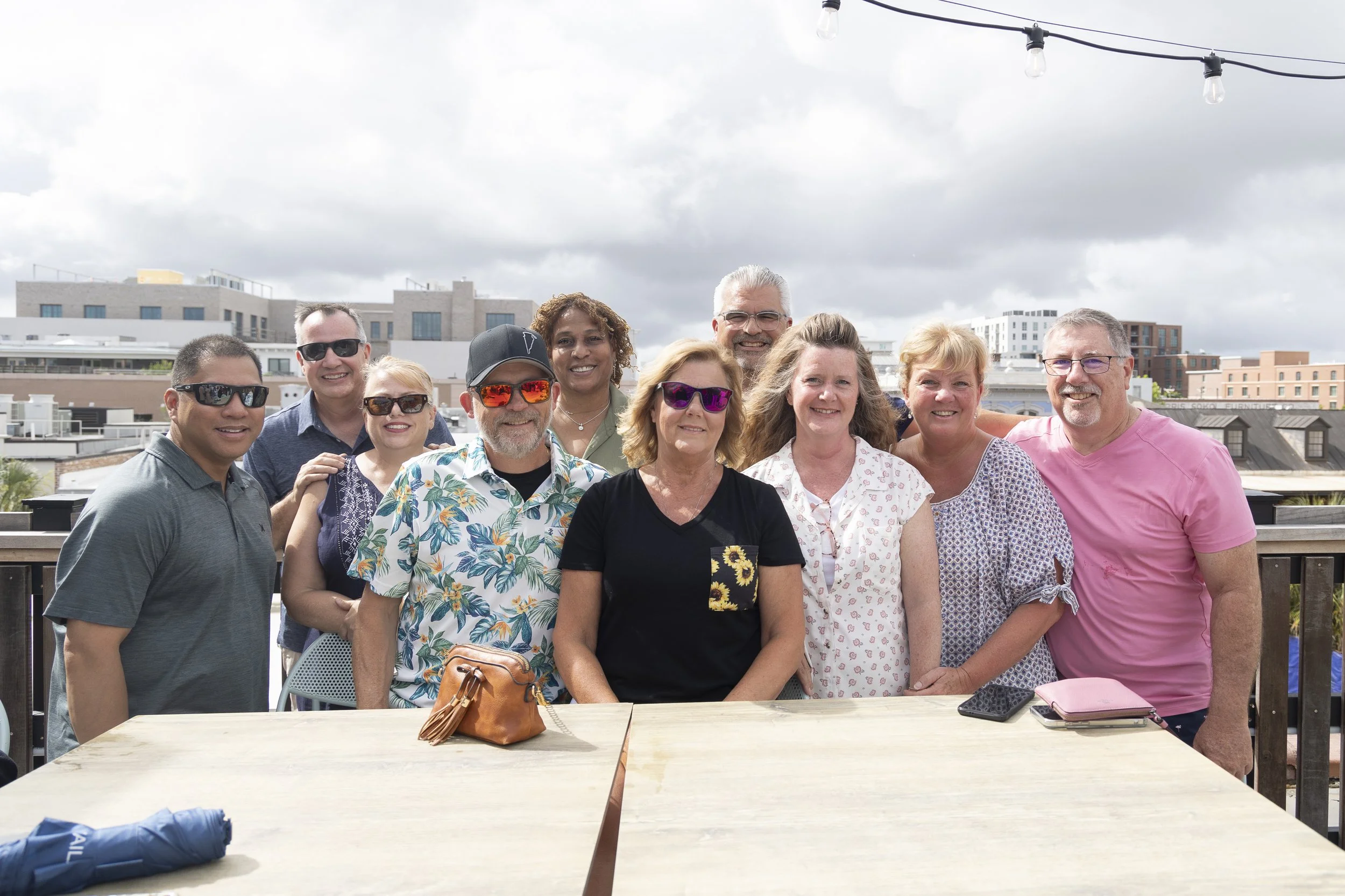 Group of ten adults standing outdoors on a balcony, smiling, with a city skyline and cloudy sky in the background, during daytime.