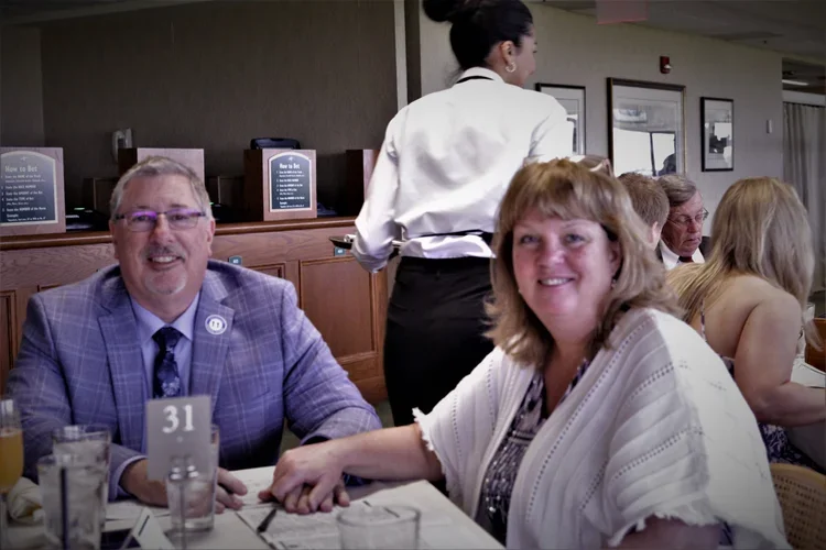 A man in a purple plaid suit and a woman in a white outfit sitting at a dining table with drinks, holding hands, in a restaurant or banquet hall.