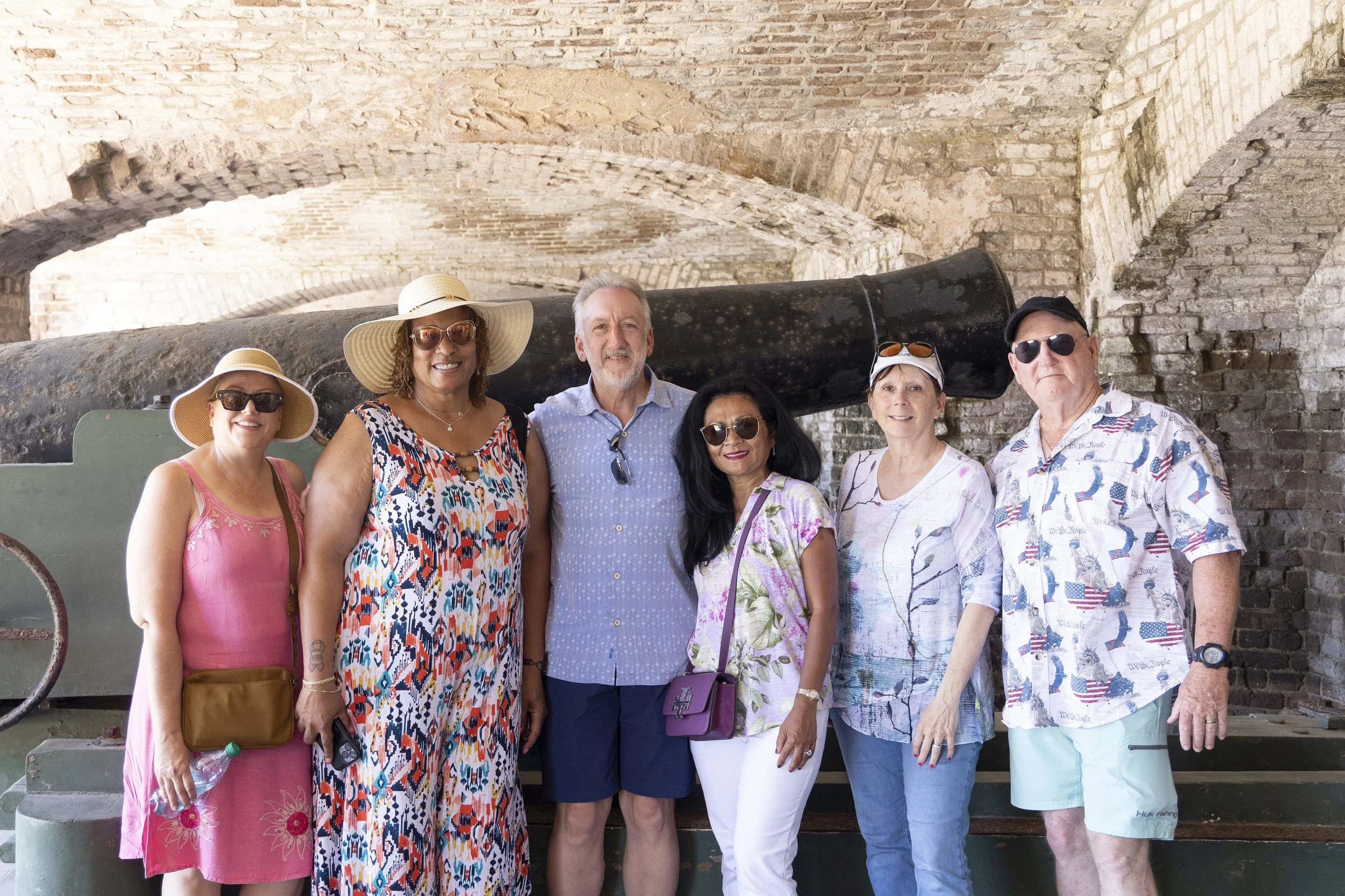 A group of six tourists standing in front of an old brick wall and a large black cannon, smiling at the camera. They are wearing summer clothes, hats, and sunglasses.