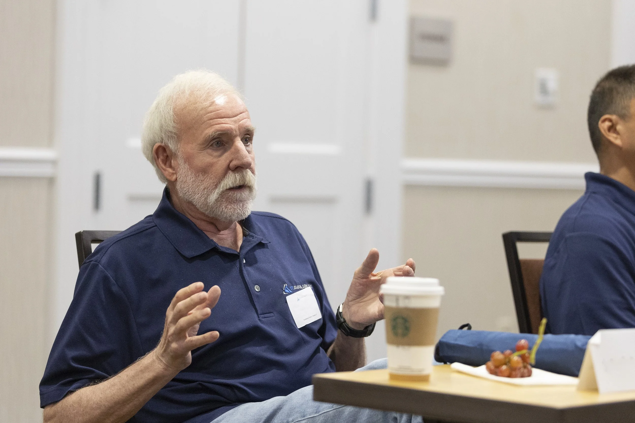 Older man with white hair and beard gesturing while seated at a table with a coffee cup, grapes, and a blue bag, in a conference room.