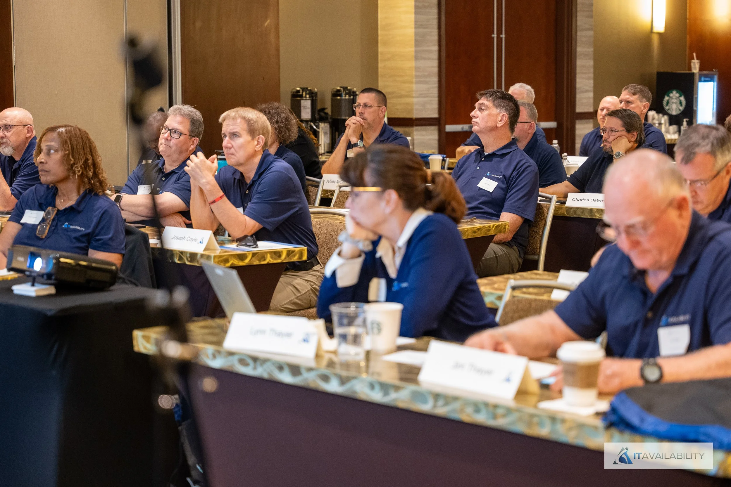 Conference attendees sitting at tables listening to a speaker in a hotel conference room, with laptops, coffee cups, and name tags in front of them.